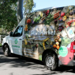 Exterior photo of a food delivery van with driver pushing a handtruck stacked with food boxes away from camera.