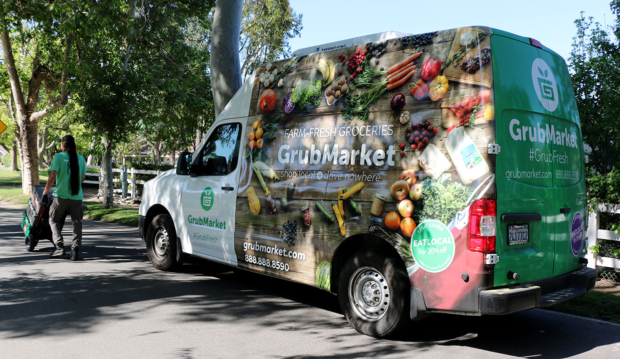 Exterior photo of a food delivery van with driver pushing a handtruck stacked with food boxes away from camera.